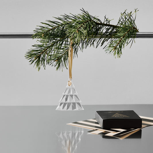 Crystal geometric pine tree ornament hanging from a pole with a pine branch on a gray surface with a white background and black gift box and wrapping paper on a reflective surface.