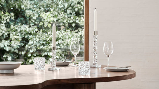 Carat collection candlesticks and votives and More collection wine glasses on a table setting with neutral wall with a window looking into a graden.
