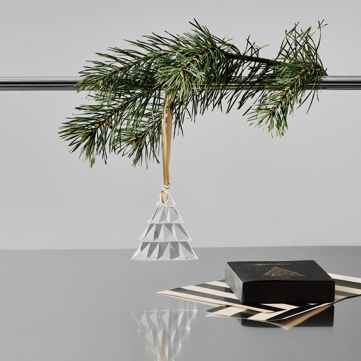 Crystal geometric pine tree ornament hanging from a pole with a pine branch on a gray surface with a white background and black gift box and wrapping paper on a reflective surface.