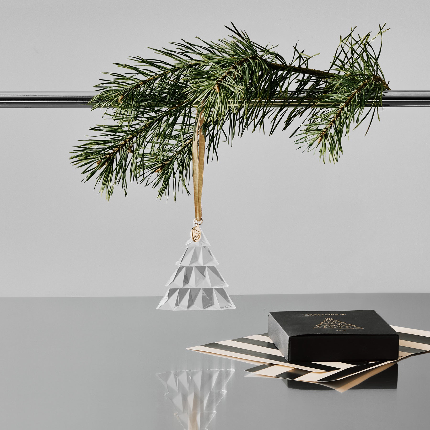 Crystal geometric pine tree ornament hanging from a pole with a pine branch on a gray surface with a white background and black gift box and wrapping paper on a reflective surface.