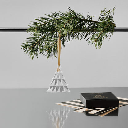 Crystal geometric pine tree ornament hanging from a pole with a pine branch on a gray surface with a white background and black gift box and wrapping paper on a reflective surface.