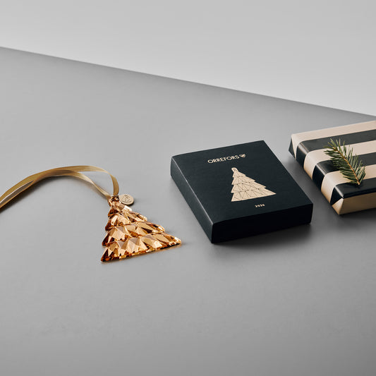 21K Gold Christmas tree ornament with a gold ribbon and a gold tag featuring the Orrefors logo on a gray surface next to a ornament gift box and gift wrapped package with a pine branch sitting on it.