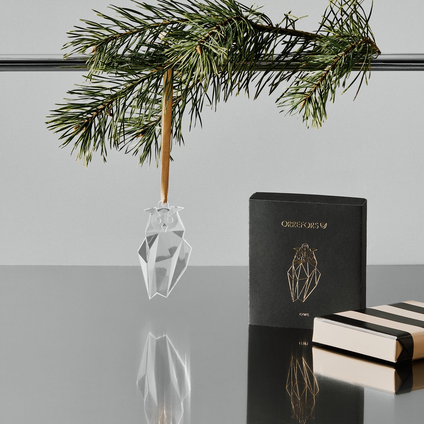 Crystal owl ornament hanging on a rod with a pine branch with a minimal background and a gift wrapped boxed sitting on a reflective surface.