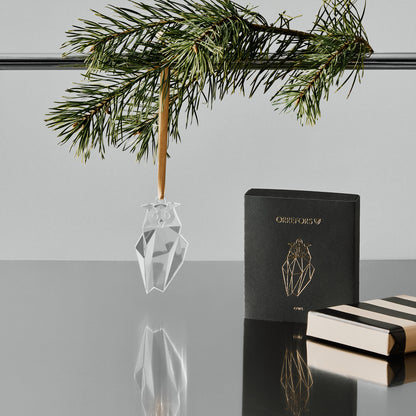 Crystal owl ornament hanging on a rod with a pine branch with a minimal background and a gift wrapped boxed sitting on a reflective surface.