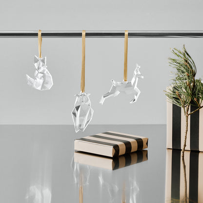 3 crystal ornaments; fox, owl, and deer hanging on a rod with a minimal background and a gift wrapped boxed and pine branch sitting on a reflective surface.