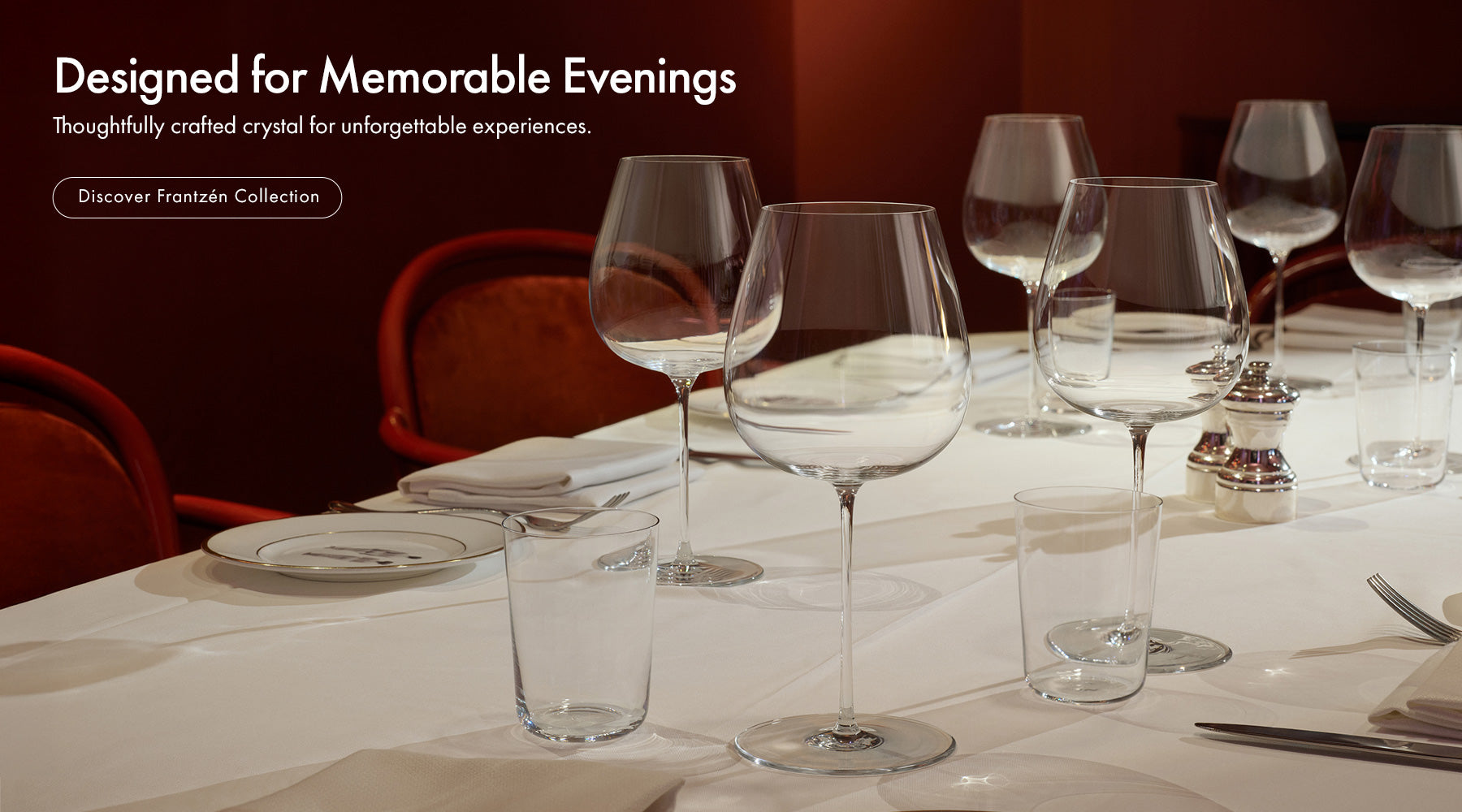 Frantzén Collection on a dining table set with clear crystal wine glasses, a tumbler, and white plates in a dimly lit dinning room setting with red chairs and a dark red wall. Text on the left reads 'Designed for Memorable Evenings' and 'Discover Frantzén Collection.