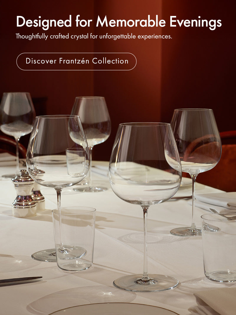 Mobile version of Frantzén Collection on a dining table set with clear crystal wine glasses, a tumbler, and white plates in a dimly lit dinning room setting with red chairs and a dark red wall. Text on the left reads 'Designed for Memorable Evenings' and 'Discover Frantzén Collection.