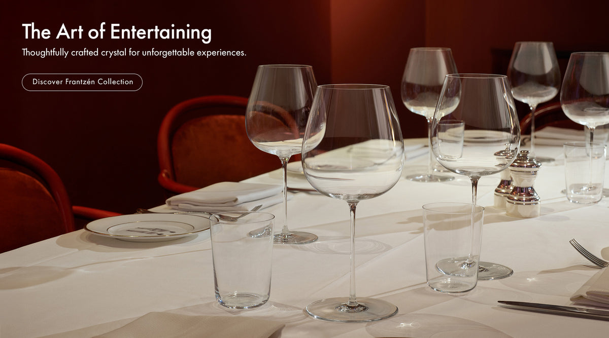 A dining table set with multiple Frantzén collection wine glasses, water glasses, and plates in a dimly lit restaurant or event space with red chairs and a dark red wall. White text on the left reads 'The Art of Entertaining' and 'Discover Frantzén Collection.
