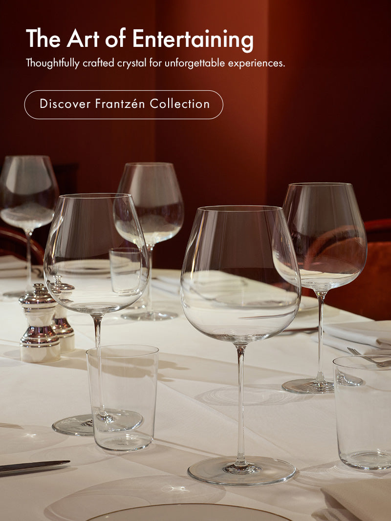 A dining table set with multiple Frantzén collection wine glasses, water glasses, and plates in a dimly lit restaurant or event space with red chairs and a dark red wall. White text on the left reads 'The Art of Entertaining' and 'Discover Frantzén Collection.