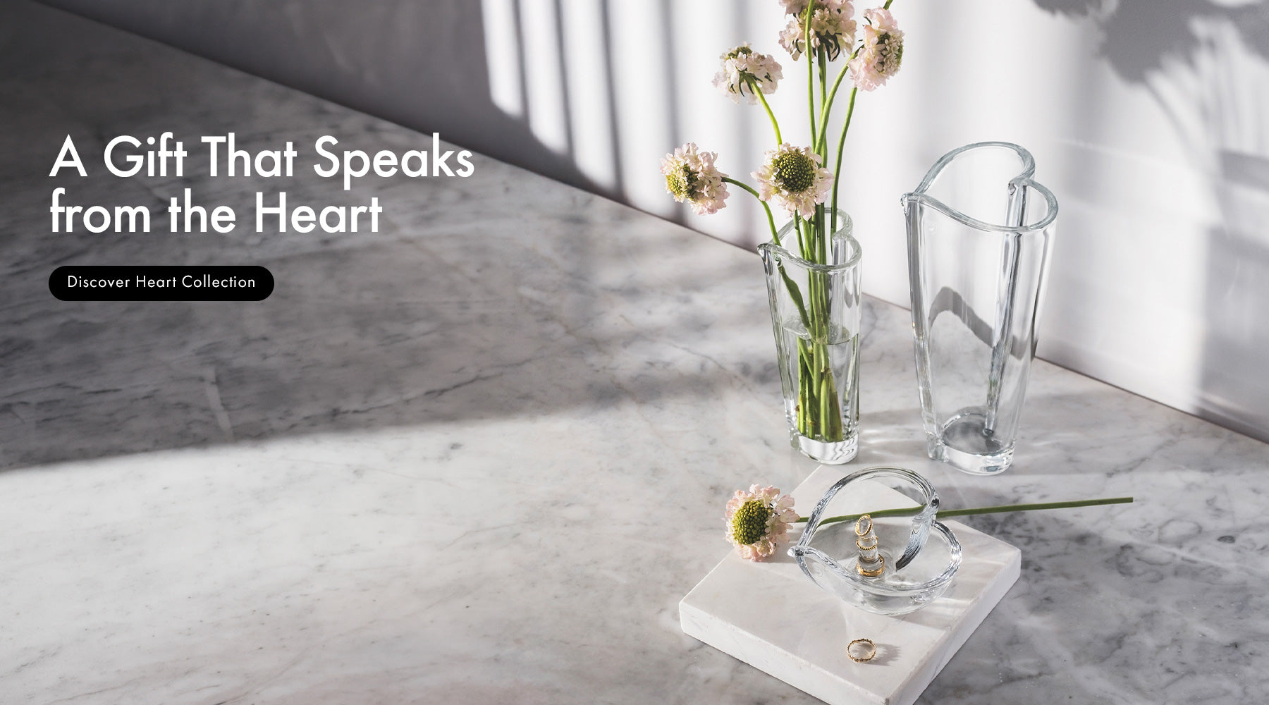 A styled interior scene on a marble surface with Heart collection heart-shaped crystal vases, in two sizes with cut flowers, and a small Heart shaped ring holder on a white marble block. Text on the left reads 'A Gift That Speaks from the Heart' and 'Discover Heart Collection.