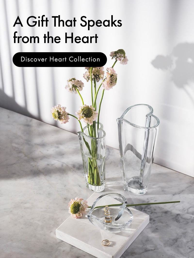 Mobile image version of a styled interior scene on a marble surface with Heart collection heart-shaped crystal vases, in two sizes with cut flowers, and a small Heart shaped ring holder on a white marble block. Text on the left reads 'A Gift That Speaks from the Heart' and 'Discover Heart Collection.