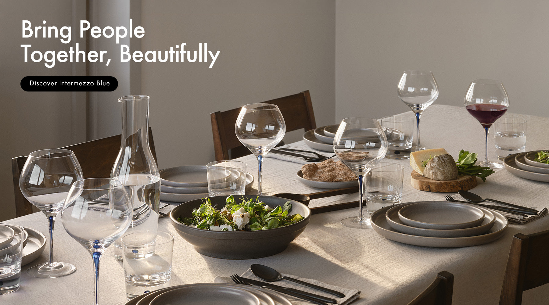A beautifully set dining table in a warm, neutral-toned room, featuring Intermezzo Blue collection wine glasses, carafe, and double old fashioned glasses. Text on the top left reads 'Bring People Together, Beautifully' and 'Discover Intermezzo Blue'.
