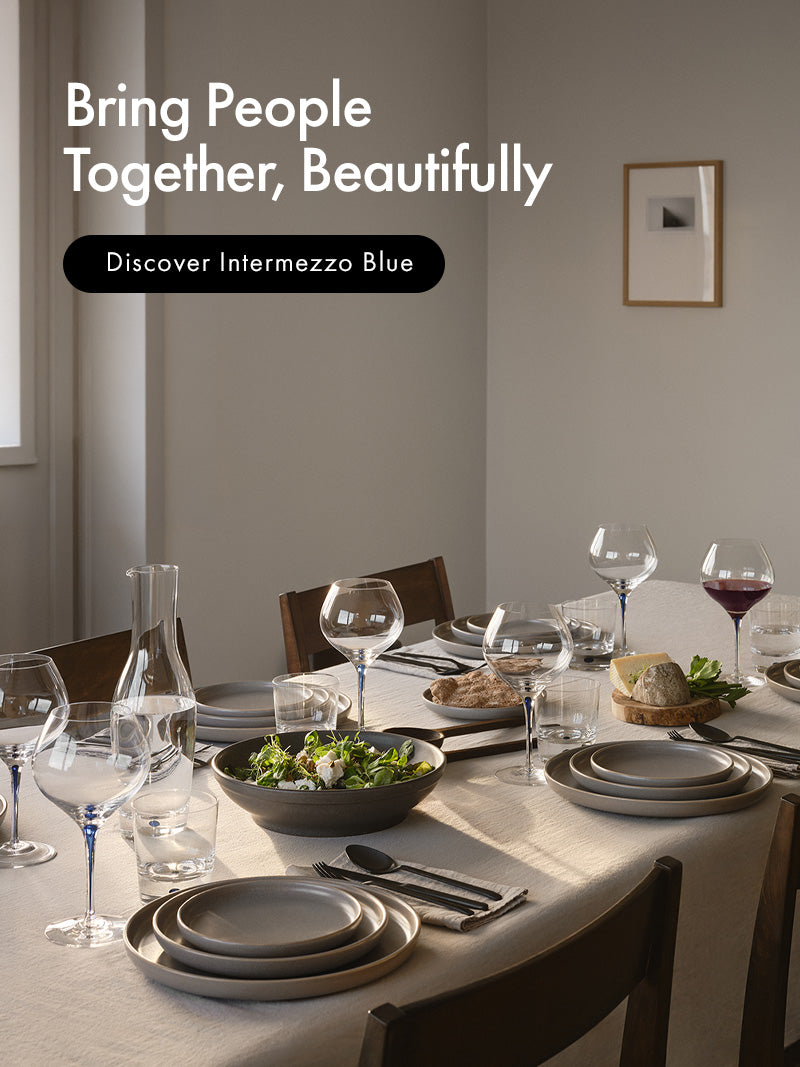 A beautifully set dining table in a warm, neutral-toned room, featuring Intermezzo Blue collection wine glasses, carafe, and double old fashioned glasses. Text on the top left reads 'Bring People Together, Beautifully' and 'Discover Intermezzo Blue'.