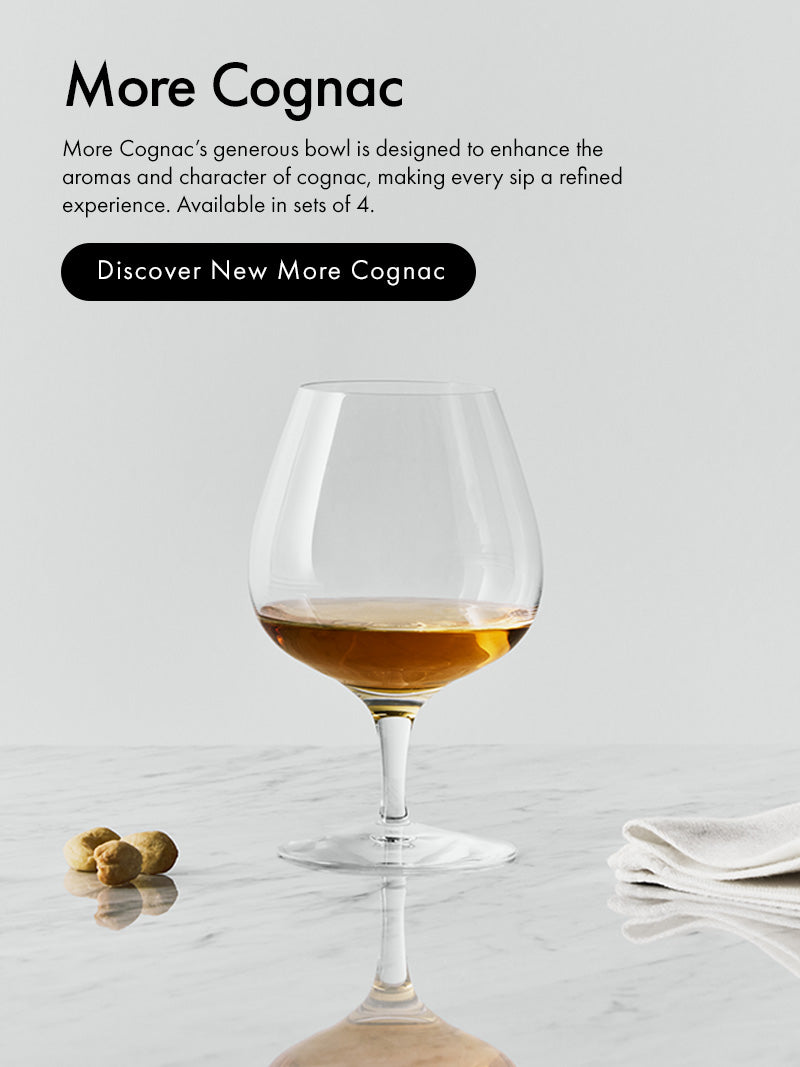 More Collection clear glass cognac snifter filled with amber cognac, set on a light marble surface with a folded white napkin and a few nuts nearby. Text on the left reads 'More Cognac' with a black button reading 'Discover New More Cognac'.