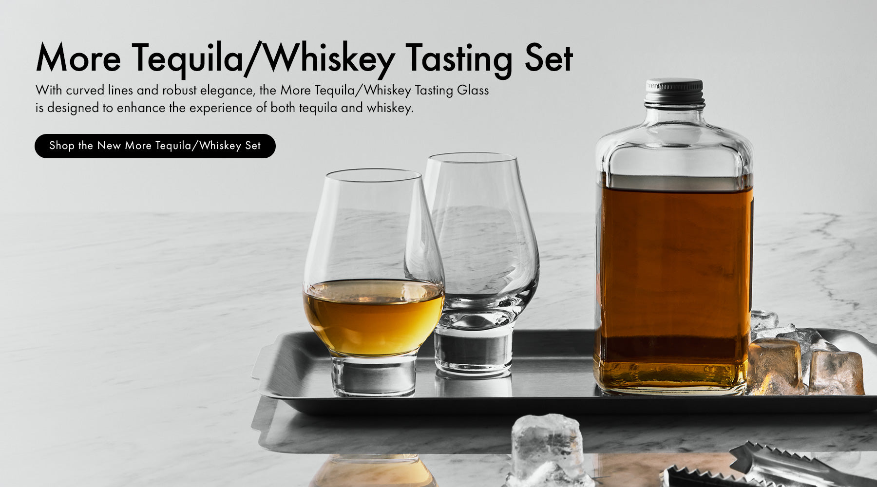 More Tequila/Whiskey Tasting Set featuring a clear glass bottle filled with amber liquor, two clear tasting glasses, and a metal tray on a light marble surface with ice cubes. Text on the left reads 'More Tequila/Whiskey Tasting Set' with a black button reading 'Shop the New More Tequila/Whiskey Set.