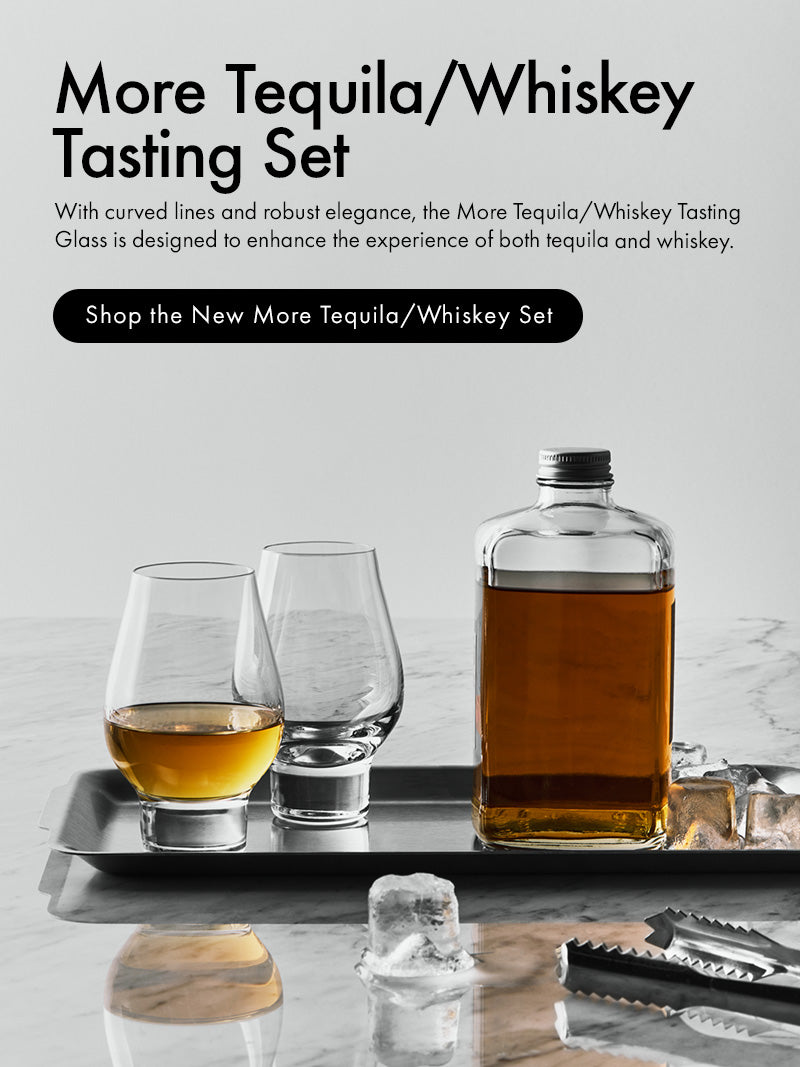 More Tequila/Whiskey Tasting Set featuring a clear glass bottle filled with amber liquor, two clear tasting glasses, and a metal tray on a light marble surface with ice cubes. Text on the left reads 'More Tequila/Whiskey Tasting Set' with a black button reading 'Shop the New More Tequila/Whiskey Set.