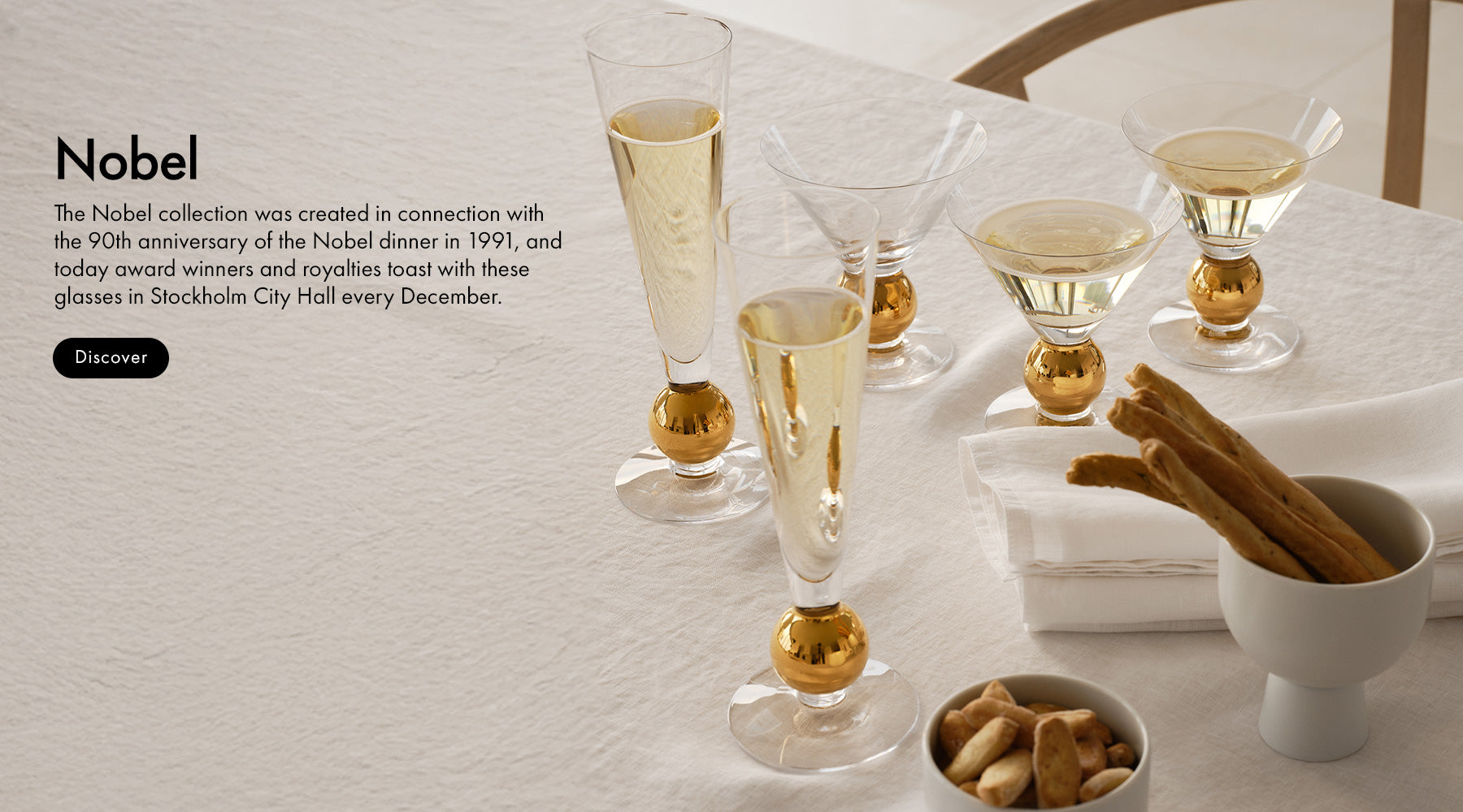 Nobel collection crystal drinkware on a  styled tabletop scene showing several clear champagne flutes with gold-colored bases, arranged on a white tablecloth beside a small bowl of cinnamon sticks and nuts. Text on the left reads 'Nobel' with a short description and a black 'Discover' button.