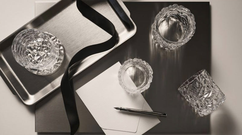 Carat collection old fashioned glass, coaster and votive candle holders on a silver tray and dark surface with a pen and note card and envelope.