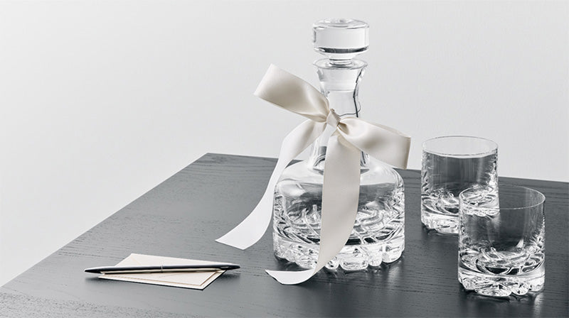 Erik Decanter Set with decanter with a ribbon tied to it and 2 double old fashioned glasses with a note card and pen on a black tabletop surface. 