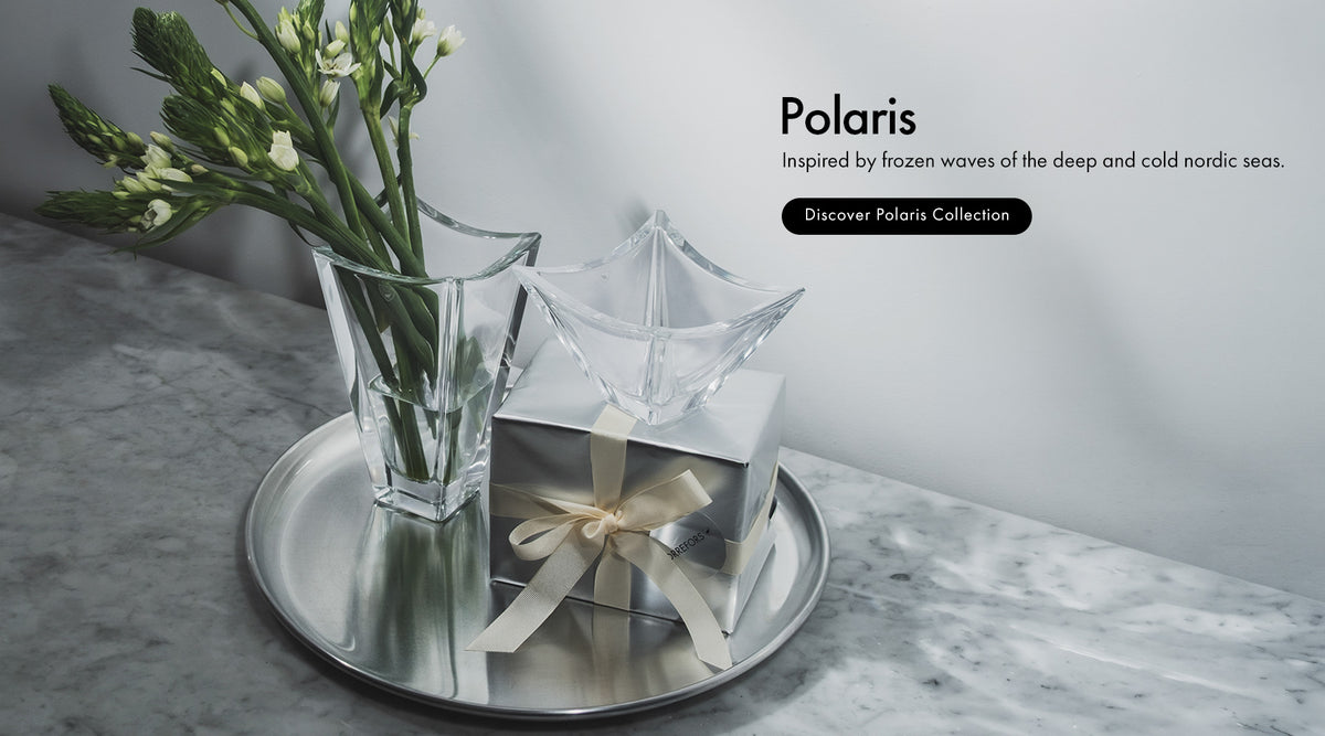 A styled product shot on a marble surface showing Polaris Collection, a clear faceted glass bowl and vase with a wrapped gift box tied with a cream ribbon, arranged on a round silver tray. Text on the right reads 'Polaris' with a tagline and 'Discover Polaris Collection'.