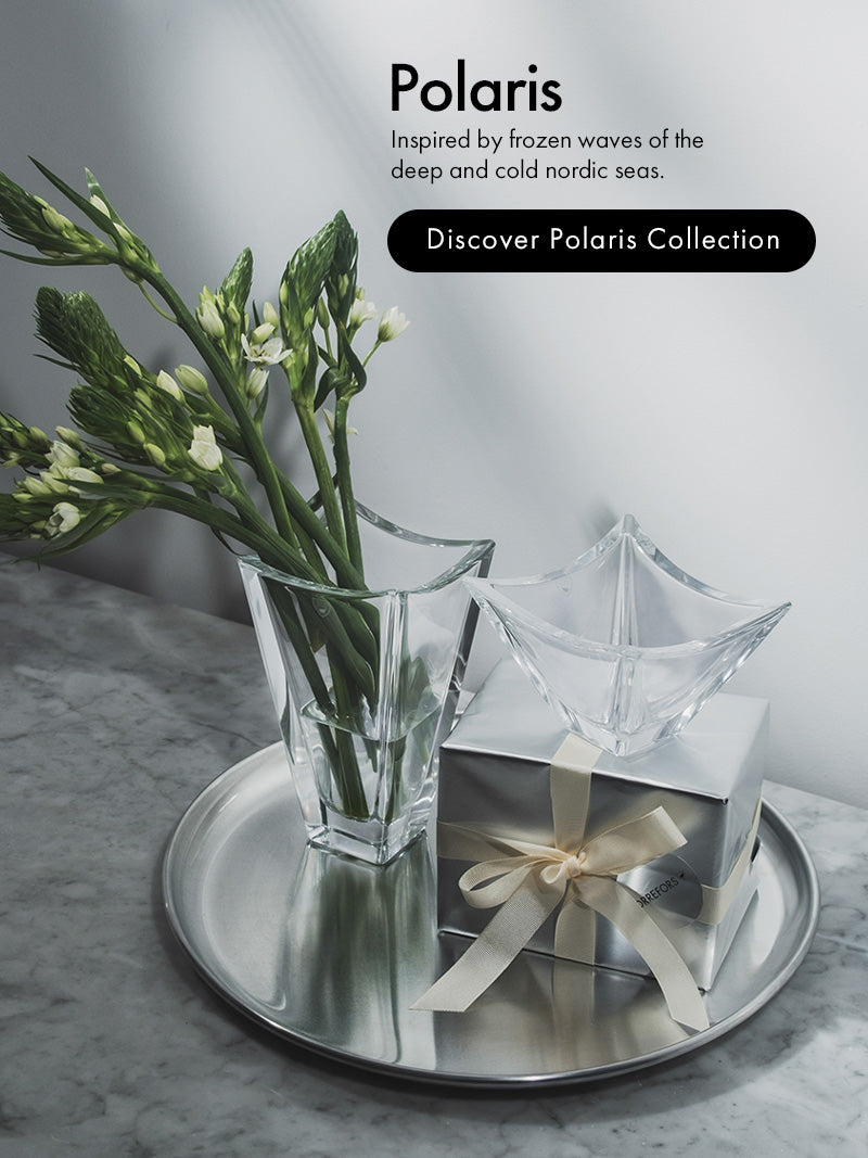 A styled product shot on a marble surface showing Polaris Collection, a clear faceted glass bowl and vase with a wrapped gift box tied with a cream ribbon, arranged on a round silver tray. Text on the right reads 'Polaris' with a tagline and 'Discover Polaris Collection'.