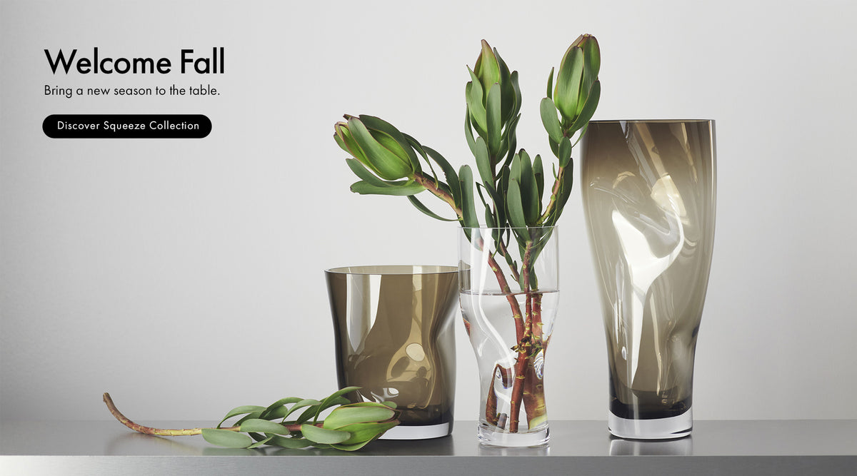 Three smoky amber glass table vases in different shapes, styled with green leaves and a water plant on a light gray background. Text on the left reads 'Welcome Fall' and 'Discover Squeeze Collection'.