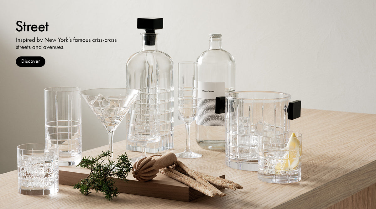 Street Collection with cut crystal grid pattern glasses, decanter, ice bucket and mineral water bottle sitting on wood surface with light background and text 'Street Inspired by New York's famous criss-cross streets and avenues.' and a Discover button.